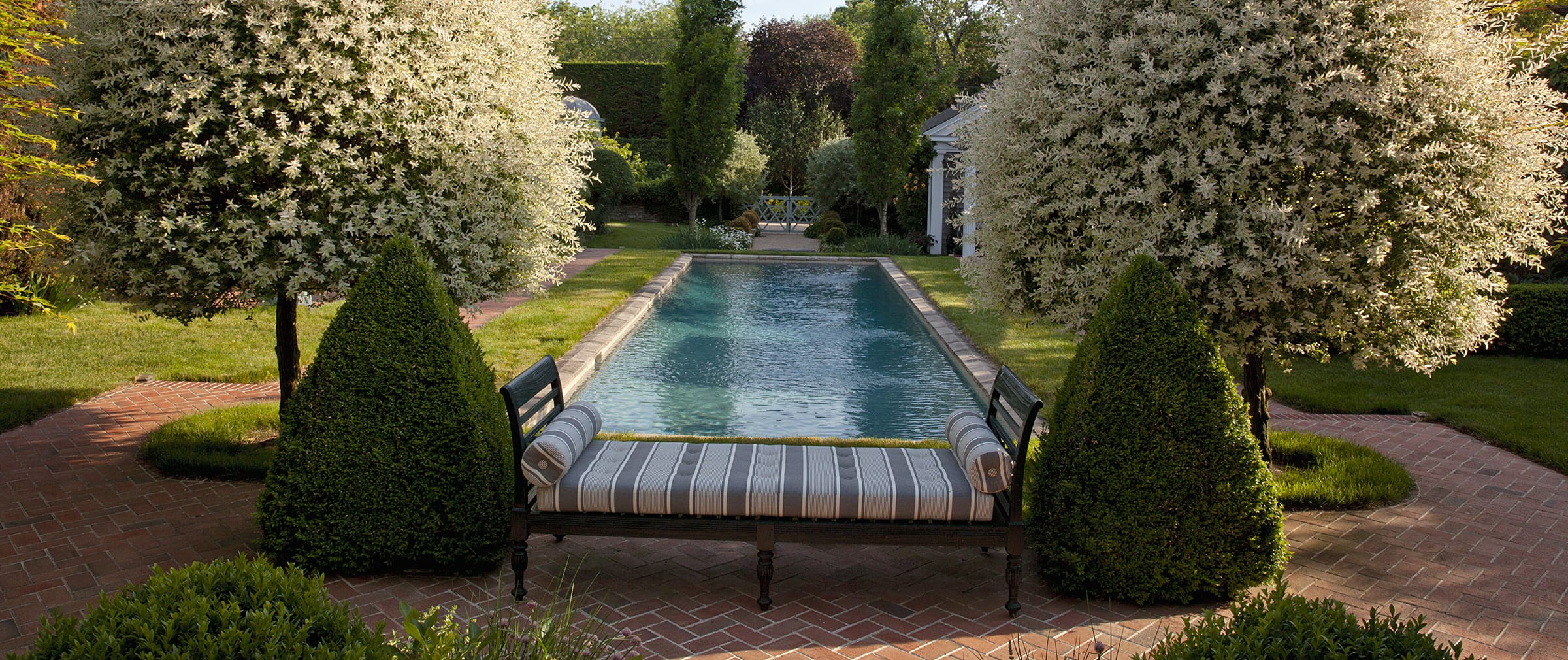 Elegant garden with a rectangular pool surrounded by lush greenery, trees, and a striped bench on a brick pathway.