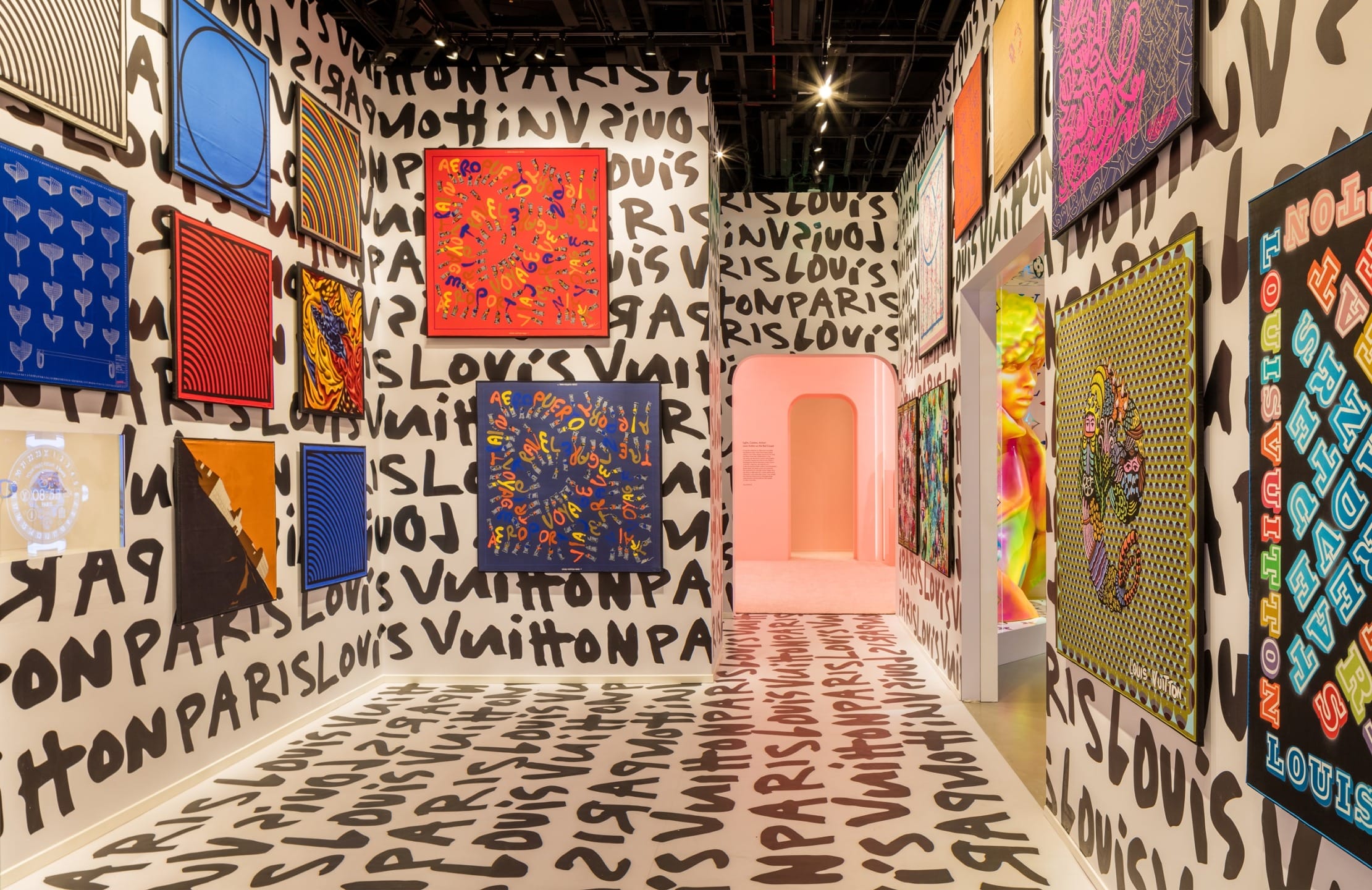 Colorful art gallery exhibit with vibrant patterns and graffiti-style text on walls and floor, leading to a pink arched doorway.