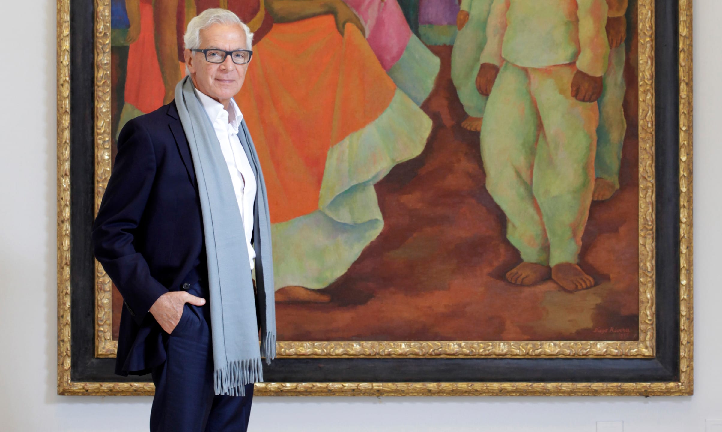 Man standing in a suit with a scarf in front of a large colorful painting displayed on a wall.