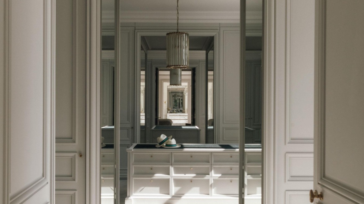 Elegant walk-in closet with mirrors, chandelier, and a hat on a countertop, featuring stylish cabinetry and natural lighting.