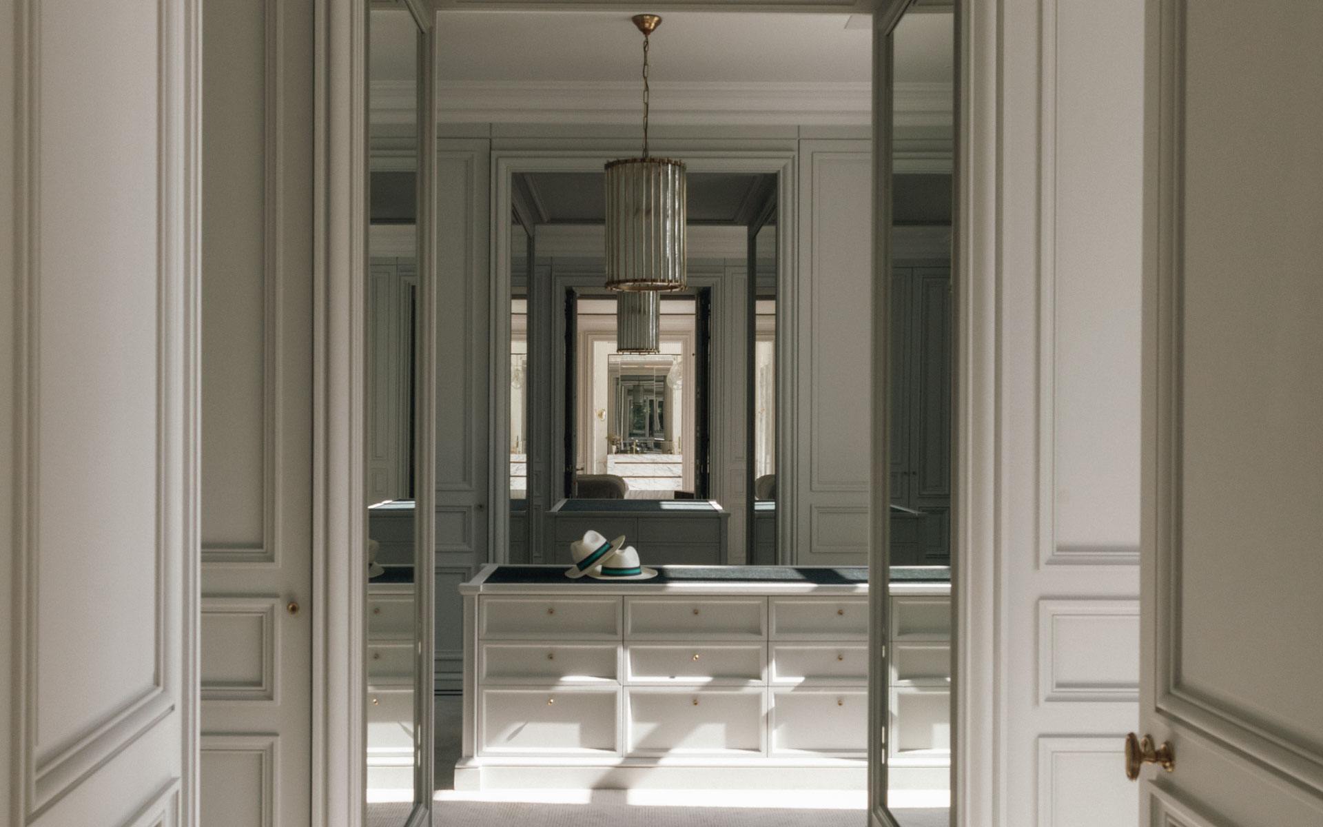 Elegant walk-in closet with mirrors, chandelier, and a hat on a countertop, featuring stylish cabinetry and natural lighting.