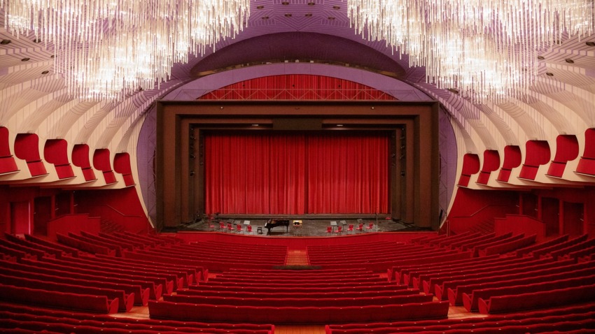 Interior of a grand theater with red seats and a stage framed by elegant chandeliers and red curtains.