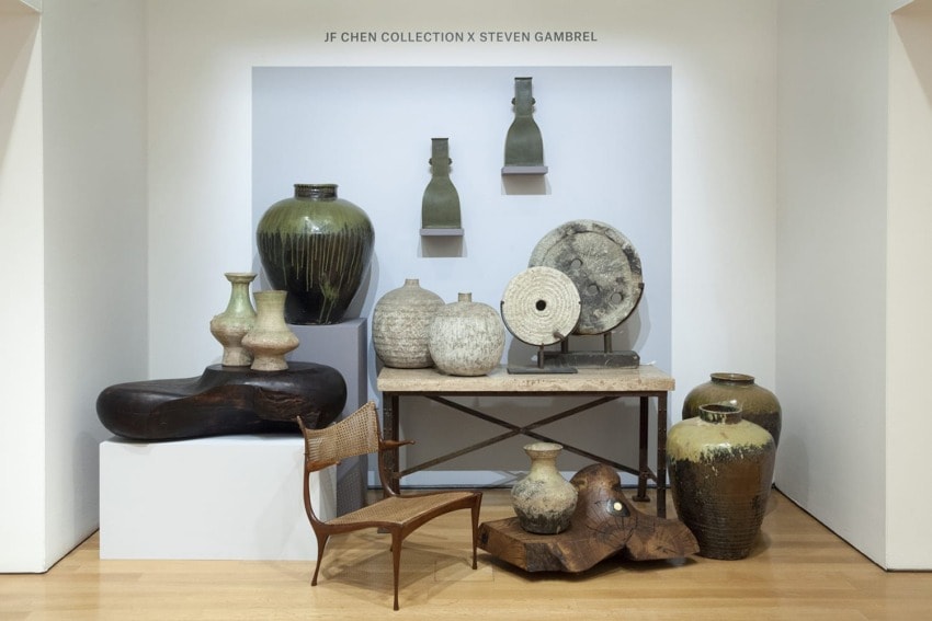 Assorted ceramics and vases displayed on pedestals and tables in a gallery setting with a wooden chair in the foreground.
