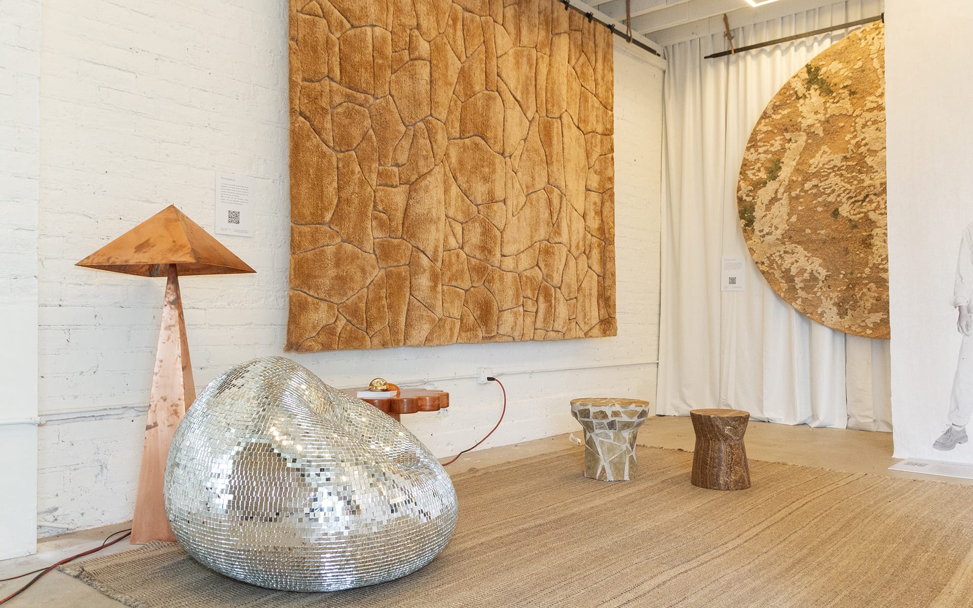 Modern art gallery with textured wall hangings, a shiny silver chair, wooden lamp, and decorative stools on a beige rug.