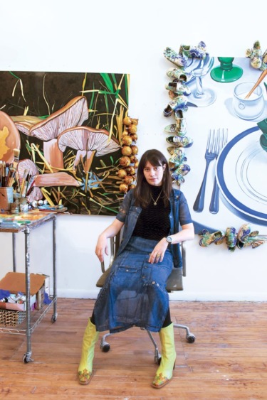 Artist seated in studio, surrounded by colorful paintings and art supplies, wearing a denim dress and yellow boots.