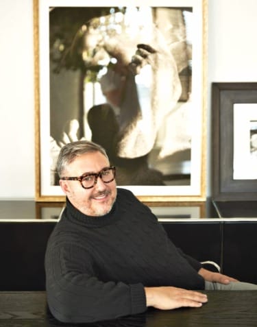 A person wearing glasses and a dark sweater sitting at a table with framed art in the background