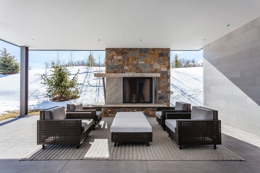Outdoor patio with wicker furniture and stone fireplace, surrounded by snow-covered landscape and evergreen trees.