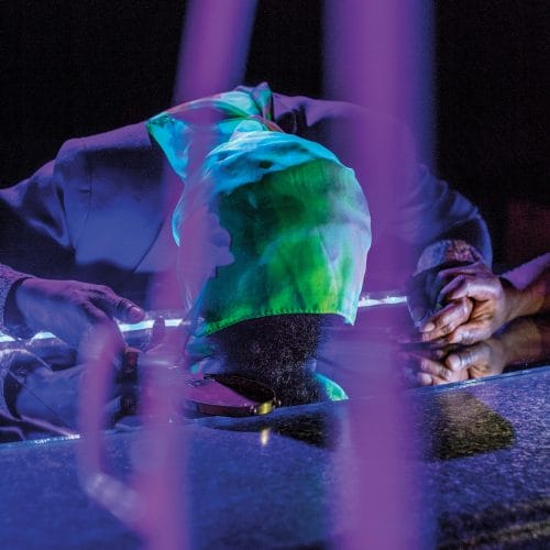 Man with colorful fabric on head leans on reflective surface in dimly lit setting, hands clasped beside him.