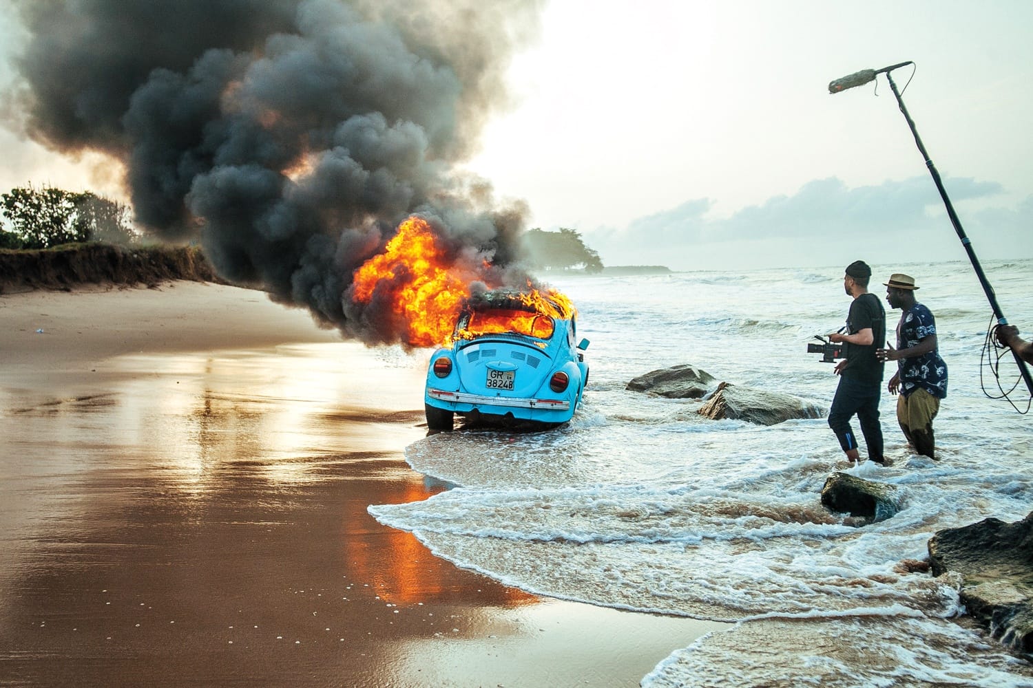 Film crew capturing a burning blue car on a beach, surrounded by waves and smoke rising into the sky.