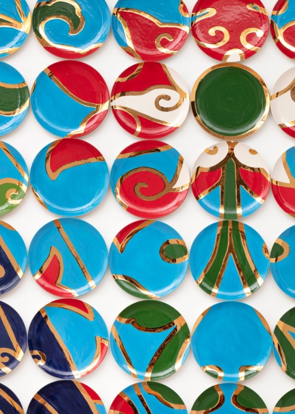 Colorful abstract ceramic plates with red, blue, green, and gold patterns arranged in a grid on a white background.