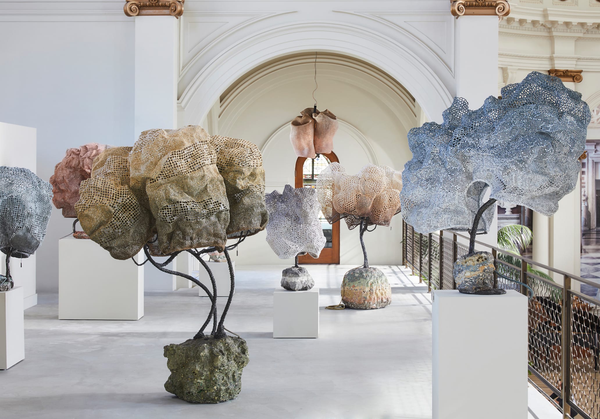 Indoor exhibition of abstract stone and mesh sculptures on pedestals in a spacious gallery with archways and natural light.