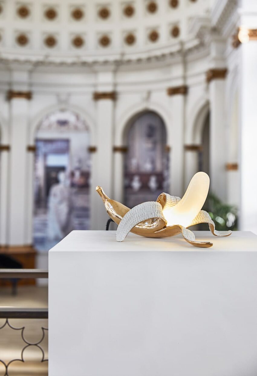 Sculpture of a peeled banana on a pedestal with a blurred historic architecture background in a gallery setting.