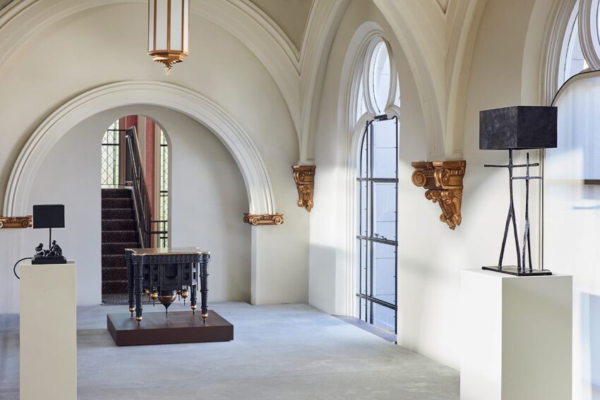 Bright room with arched windows, showcasing modern sculptures on pedestals and a decorative table in the center.