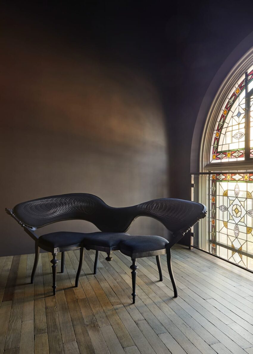 Modern black sculptural bench near an ornate stained glass window in a dimly lit room with wooden floor.