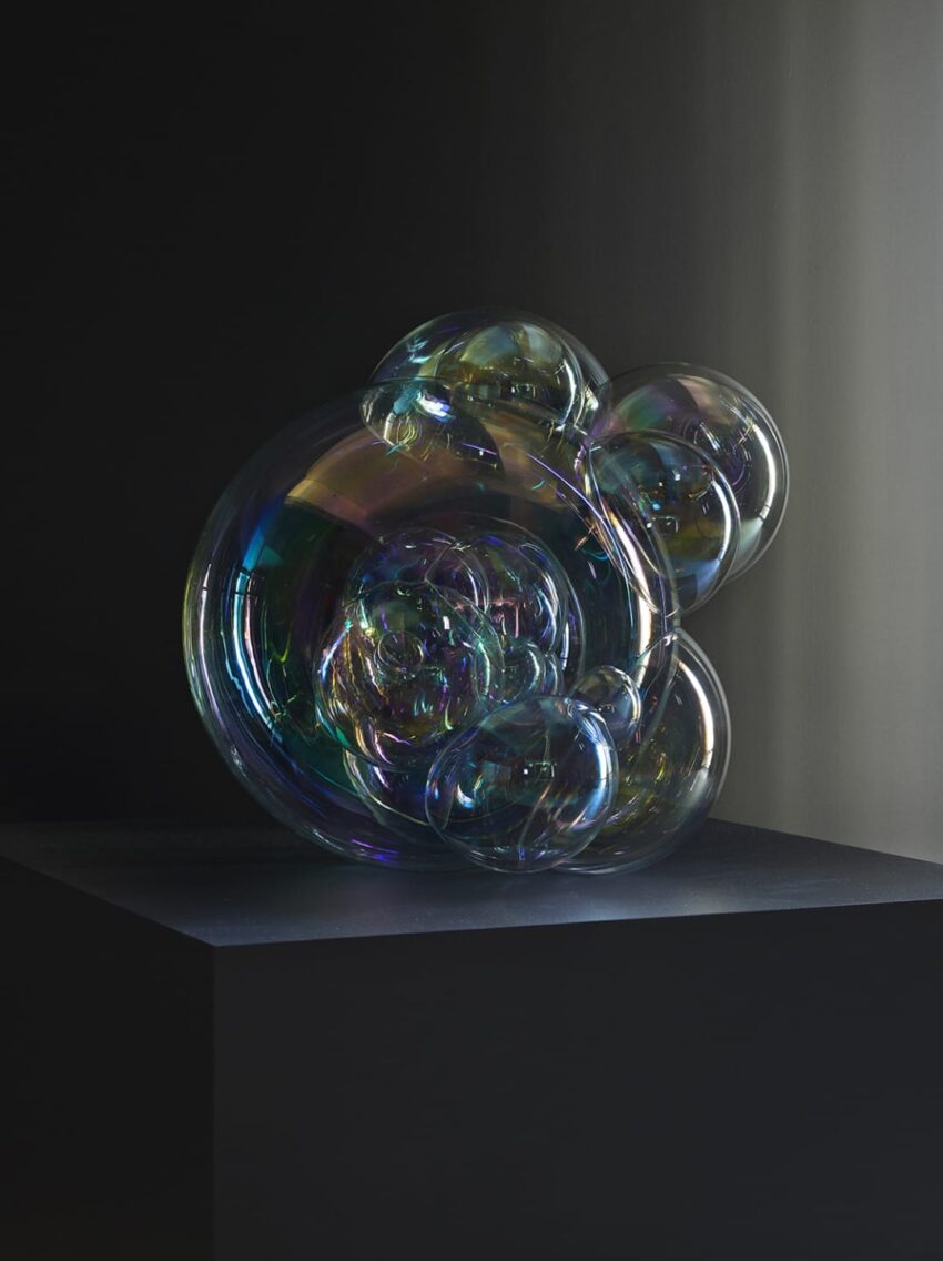 Colorful glass sculpture with bubble-like shapes displayed on a dark pedestal, illuminated by soft lighting.