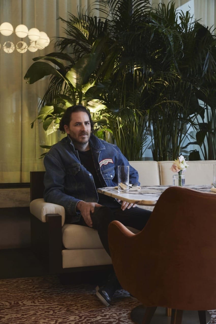 Person in denim jacket sitting at a table in a stylish, plant-filled indoor setting with velvet chairs and soft lighting.