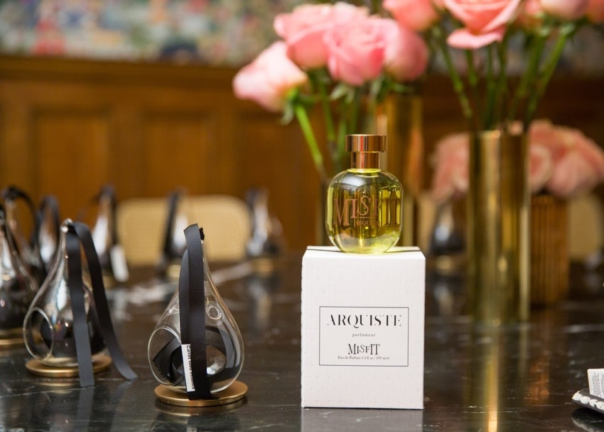 Arquiste Misfit perfume bottle on a box with pink flowers in the background on a dark table.