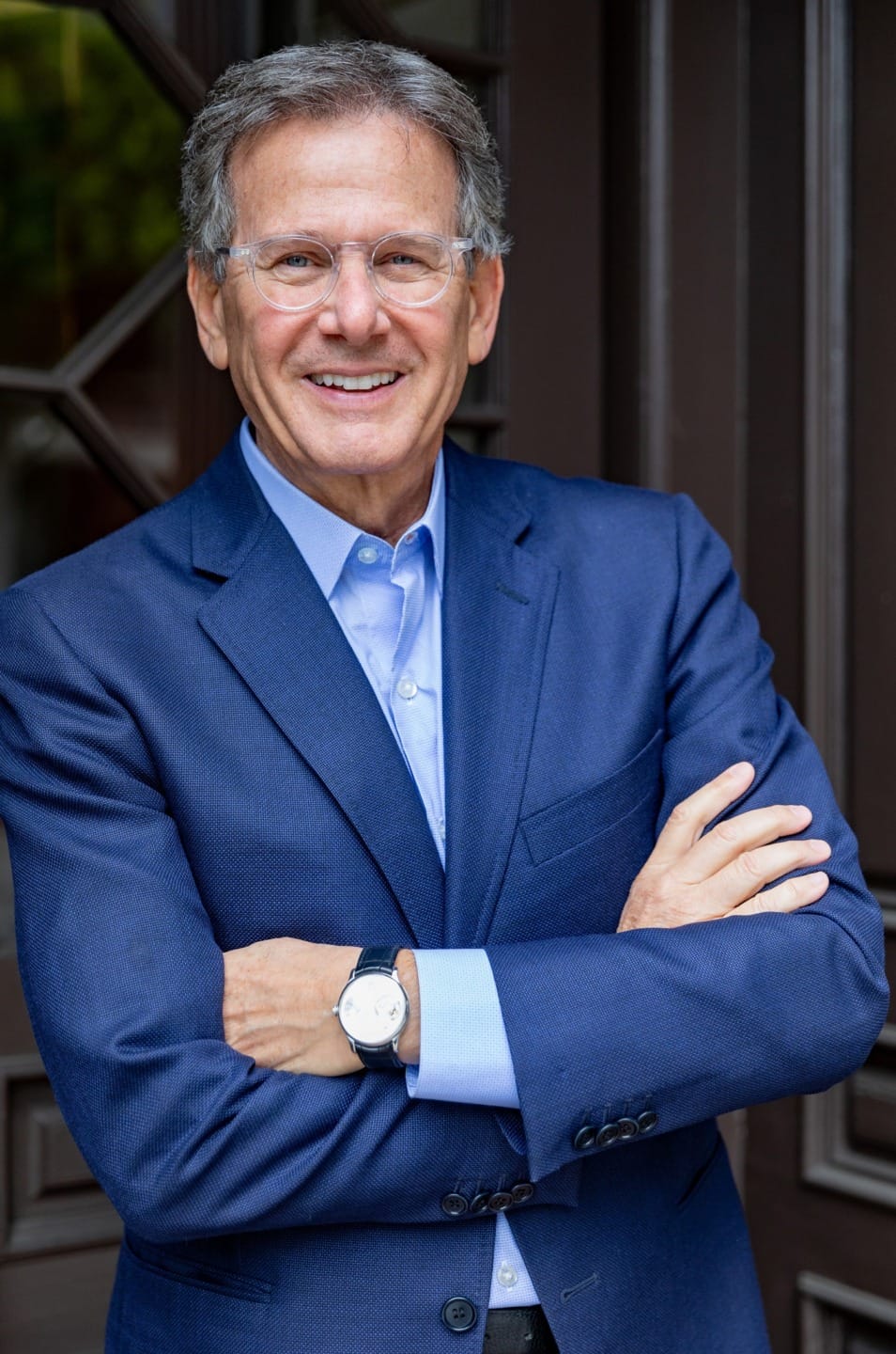Man in a blue suit and glasses smiling with arms crossed, standing in front of a wooden door.