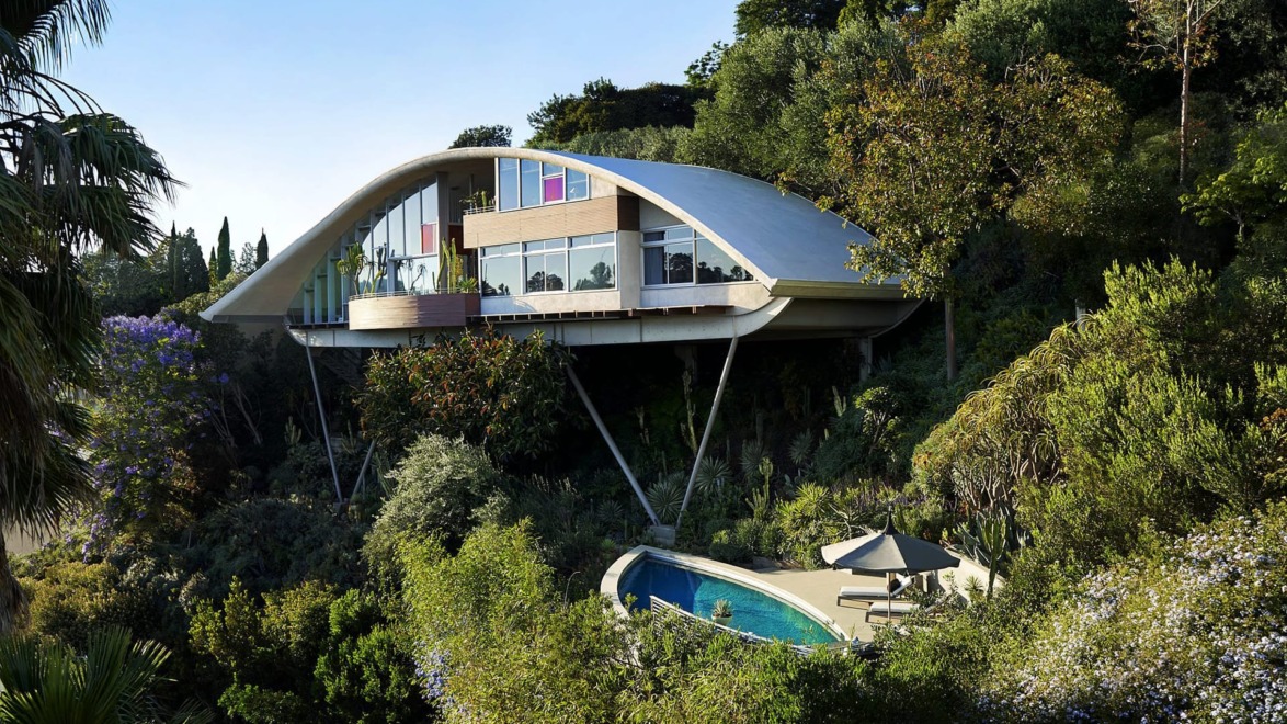 Futuristic house with arched roof, large windows, surrounded by trees, and an outdoor pool visible in a lush setting.
