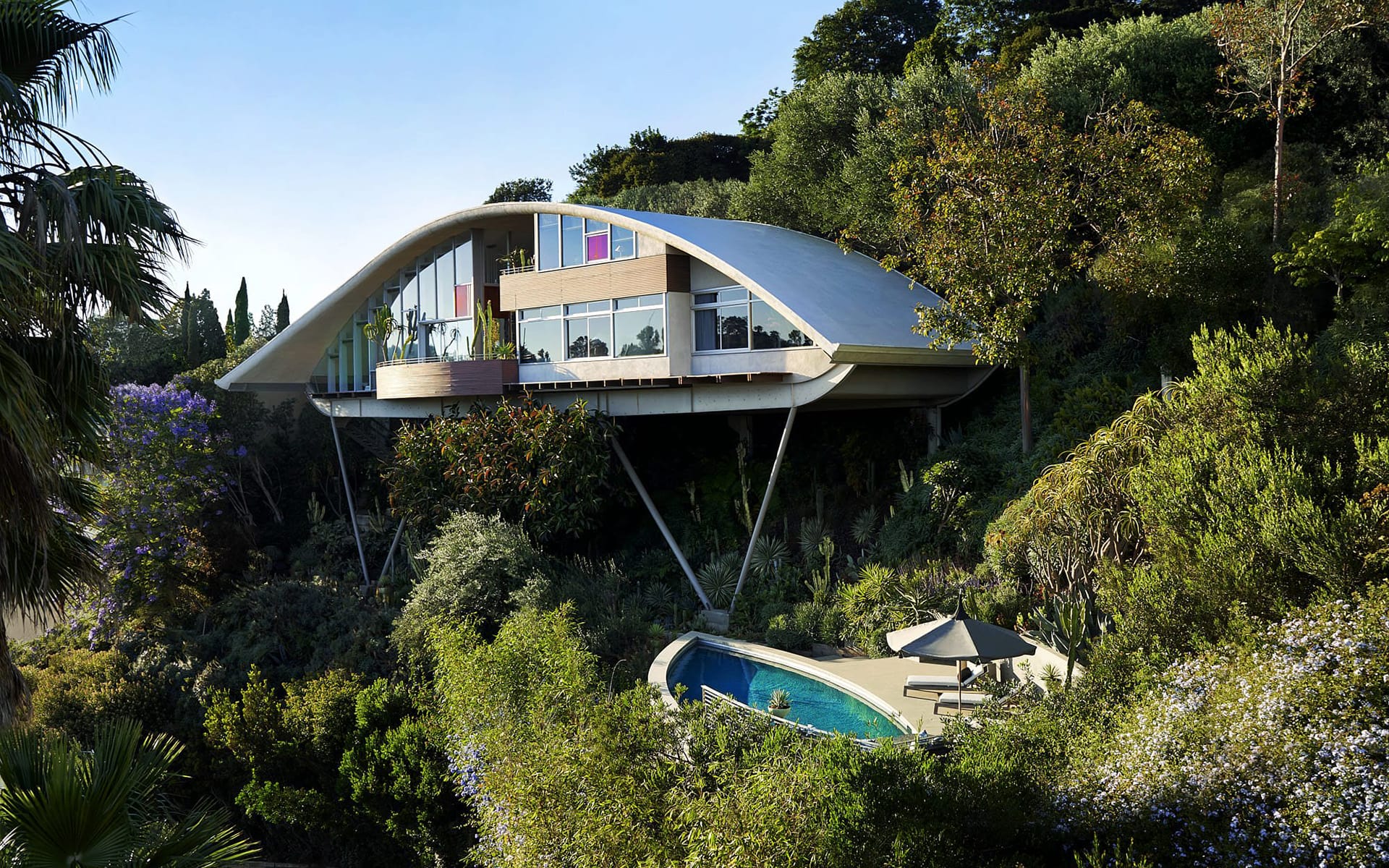 Futuristic house with arched roof, large windows, surrounded by trees, and an outdoor pool visible in a lush setting.