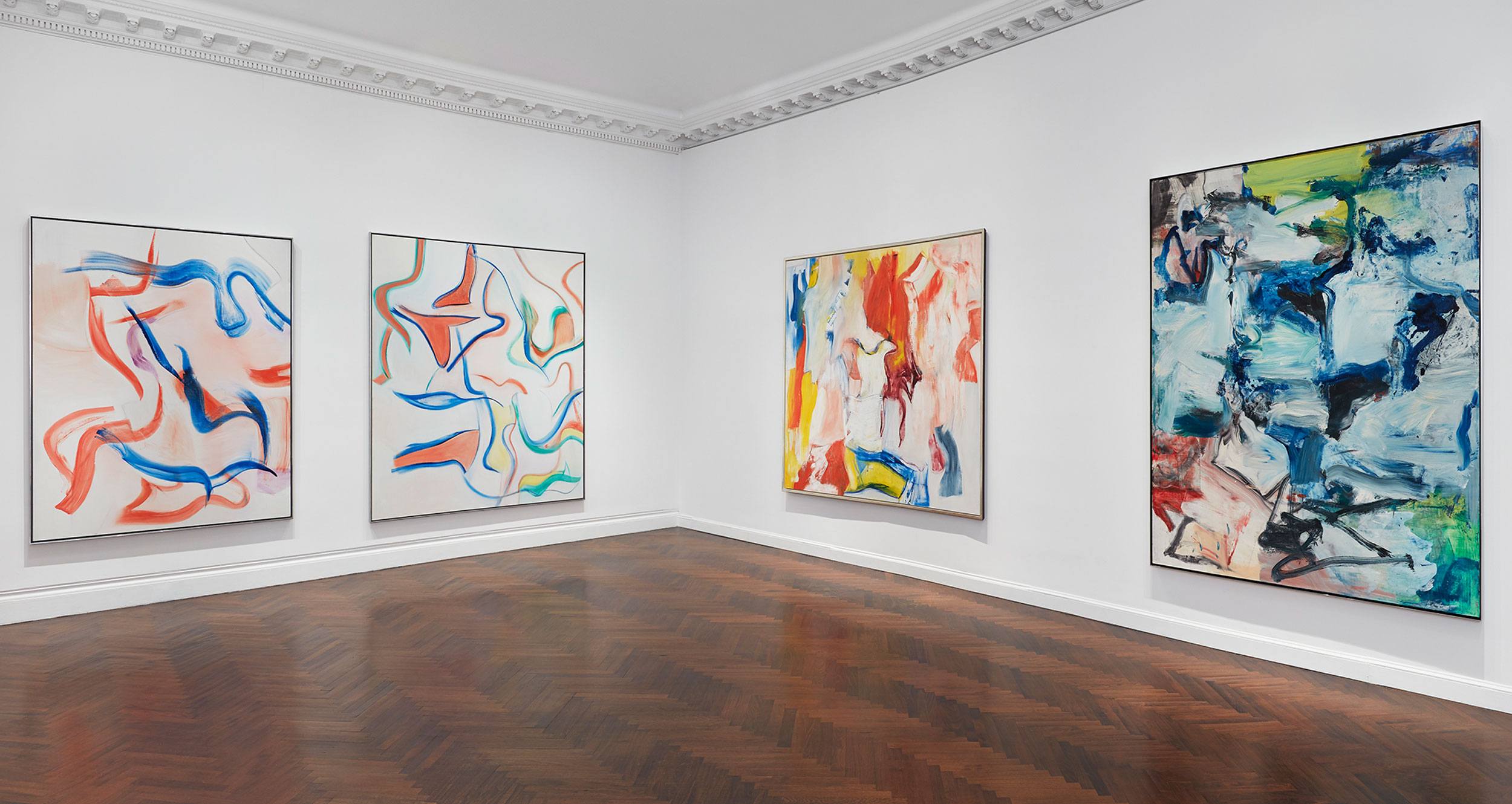 Art gallery room displaying four abstract paintings with vibrant colors on a polished wooden floor and white walls.