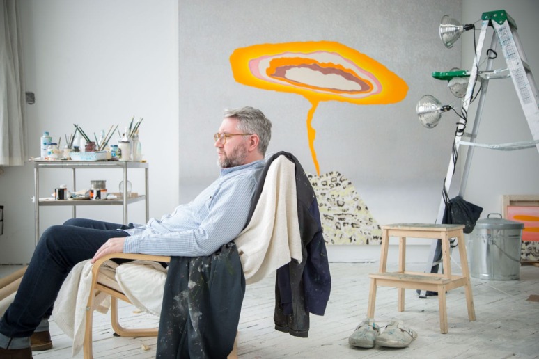 Artist sitting in a studio with painting supplies and a colorful artwork on the wall behind him