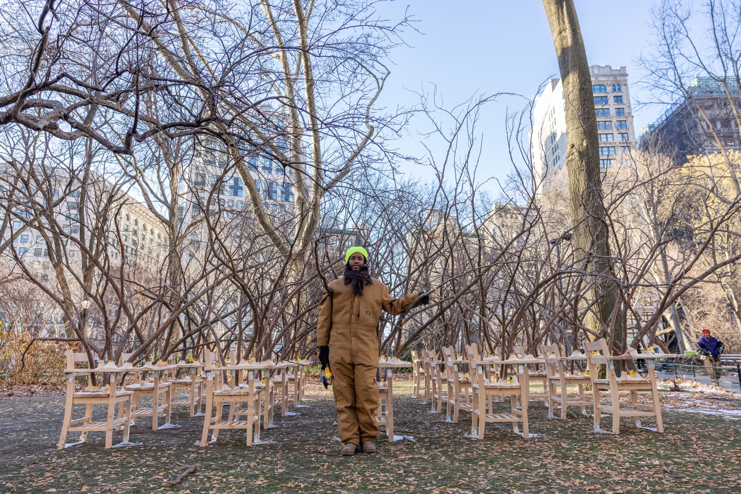 Person in brown coveralls and green hat stands in outdoor art installation with curved branches and wooden chairs, winter scene.
