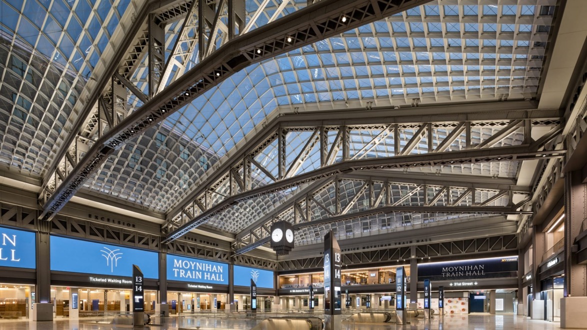 Interior view of Moynihan Train Hall featuring a spacious glass ceiling, information displays, and an iconic clock.