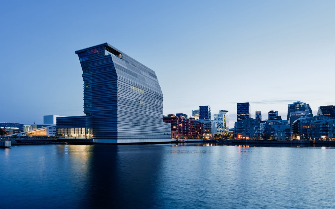 Modern waterside building with city skyline in the background during twilight, featuring unique architecture and reflective water.