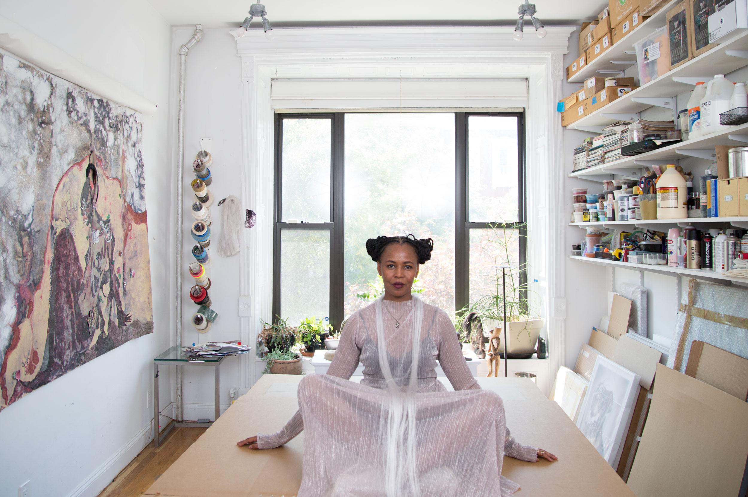 Person in sheer outfit sitting cross-legged on table in art studio with large window and shelves filled with supplies.