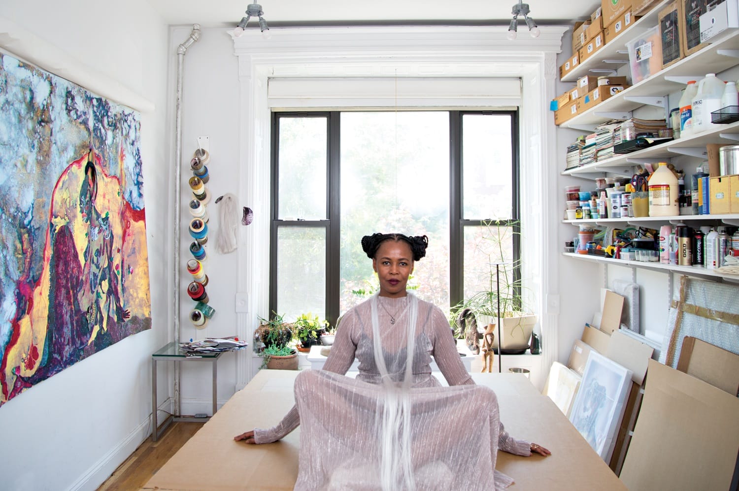 Artist sitting in a bright studio with colorful artwork on the wall and shelves filled with art supplies, looking at the camera.