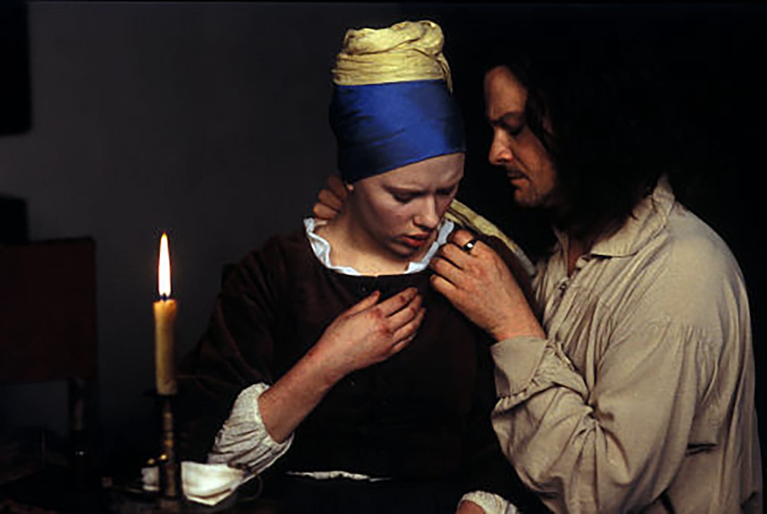 Painter examining woman's clothing by candlelight in dimly lit room, with both figures focused on the fabric.