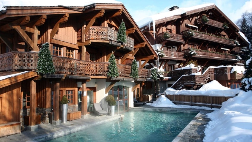 Wooden chalet with snow-covered roof and balconies, next to a heated outdoor pool, surrounded by a snowy landscape.