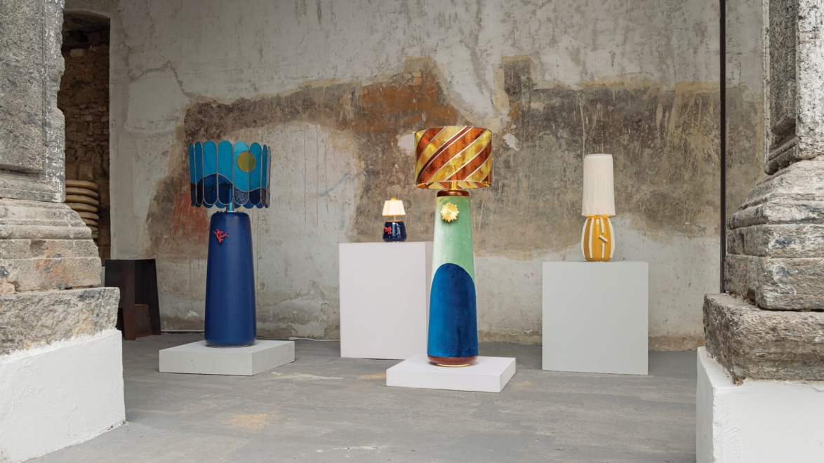 Colorful, artistic lamps displayed on pedestals against a textured, rustic wall in a gallery setting.