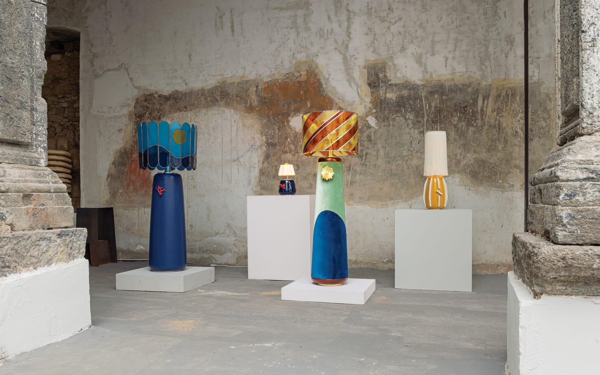 Colorful, artistic lamps displayed on pedestals against a textured, rustic wall in a gallery setting.