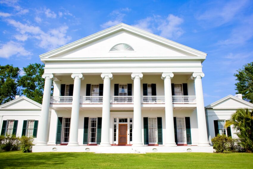 Large white mansion with tall columns and green shutters against a clear blue sky.