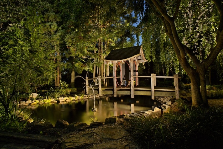 Japanese garden with a wooden bridge over a pond, surrounded by lush trees, softly lit in the evening.