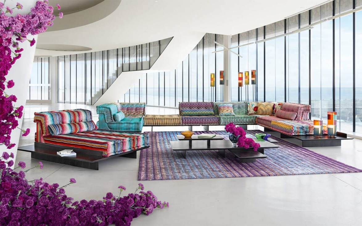 Modern living room with colorful striped couches, large windows, purple flowers, and vibrant decor on a patterned rug.