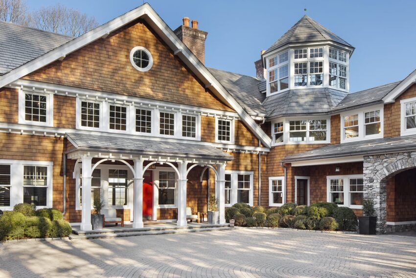 Large, elegant house with brown siding, white trim, and circular driveway under a clear blue sky.
