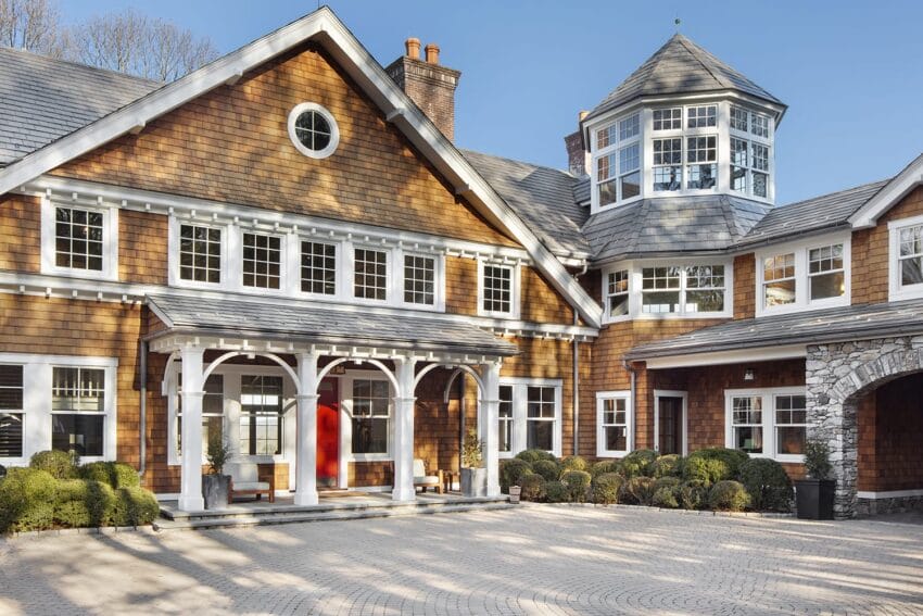 Large, elegant house with brown siding, white trim, and circular driveway under a clear blue sky.