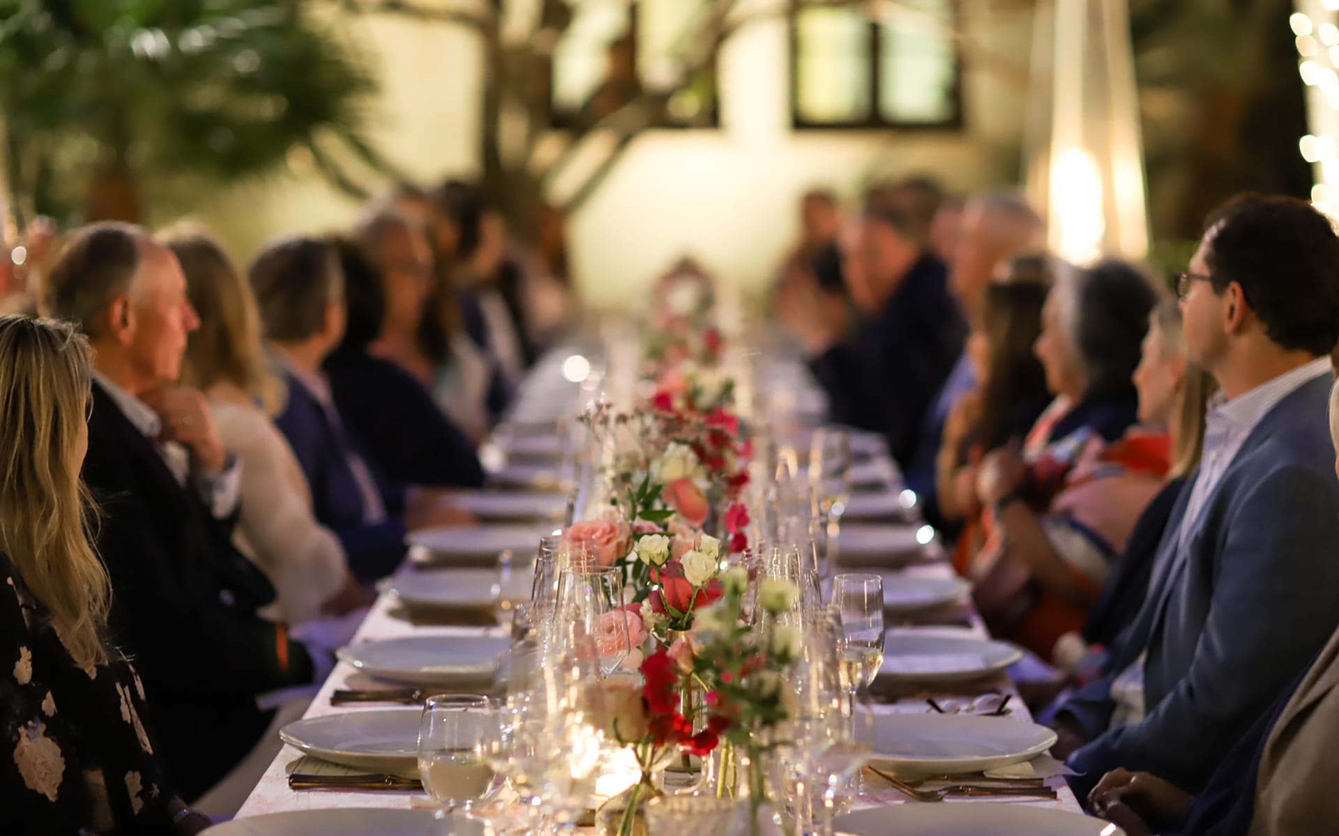People sitting at a long, elegantly set dinner table with flowers and candles, engaged in conversation during an evening event.