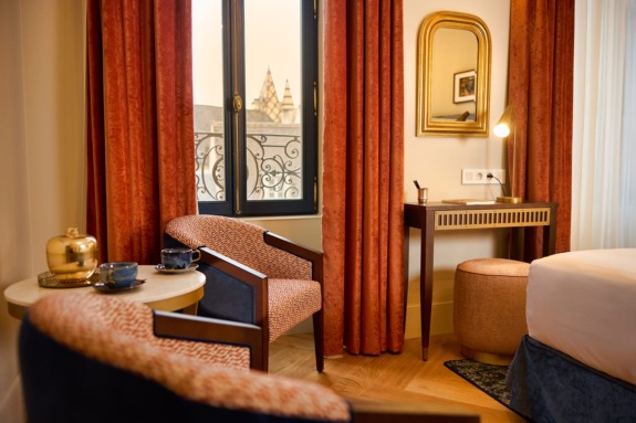 Elegant hotel room with two chairs, a table set for tea, a writing desk, and large window with city view in background.