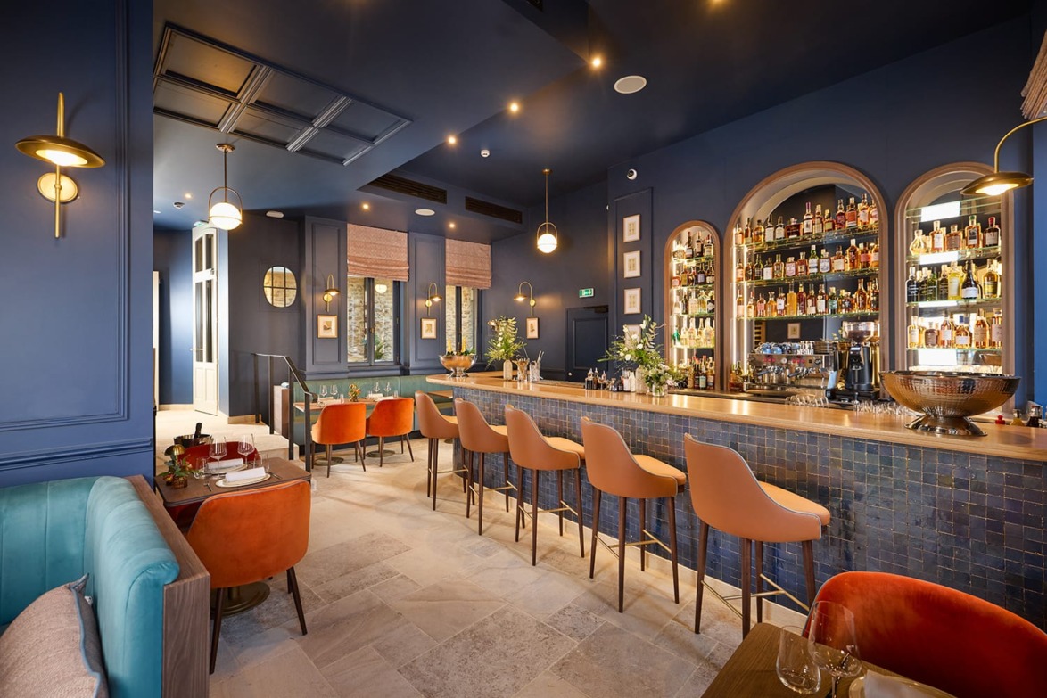 Stylish modern bar interior with blue walls, a long counter, orange stools, and shelves filled with bottles.
