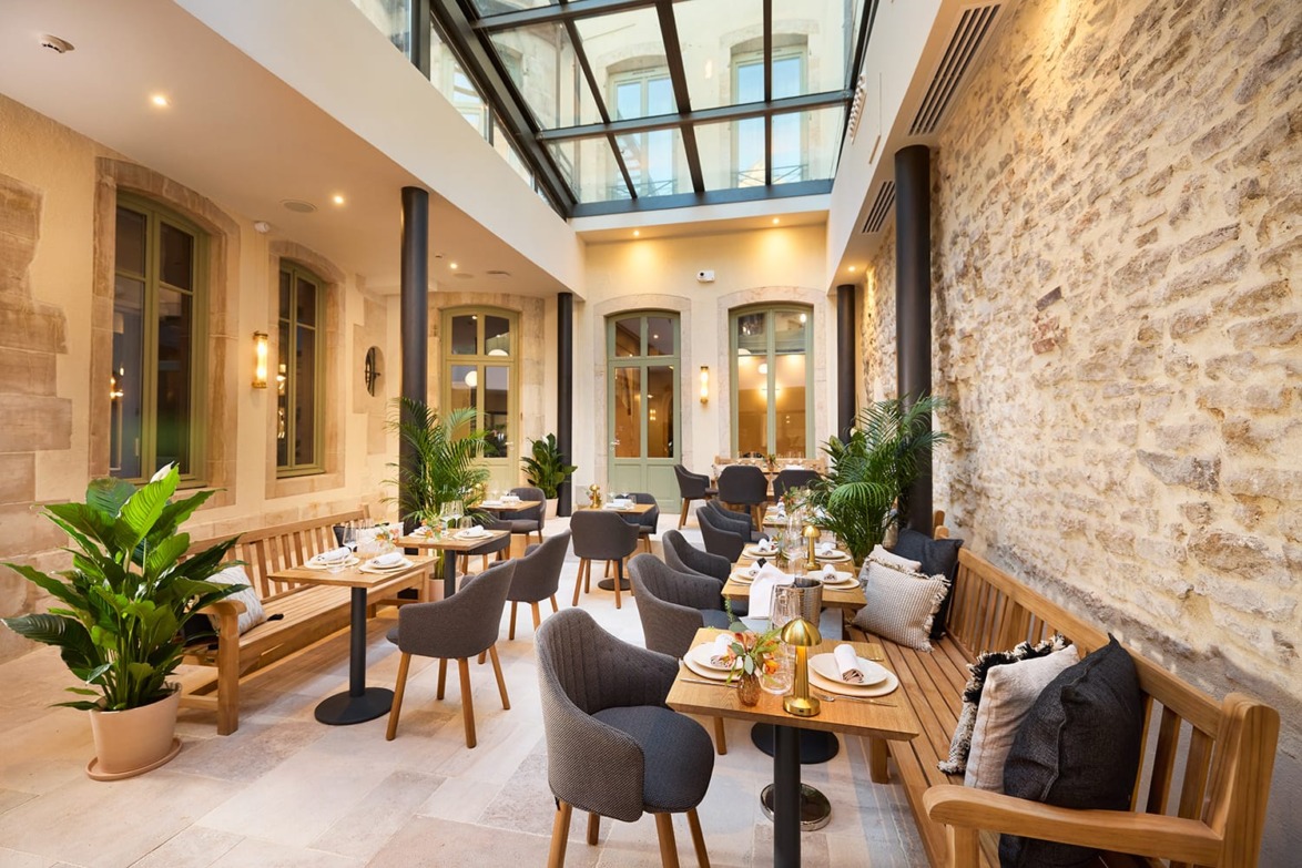 Bright indoor cafe with wooden furniture, potted plants, and natural light streaming through a glass ceiling.