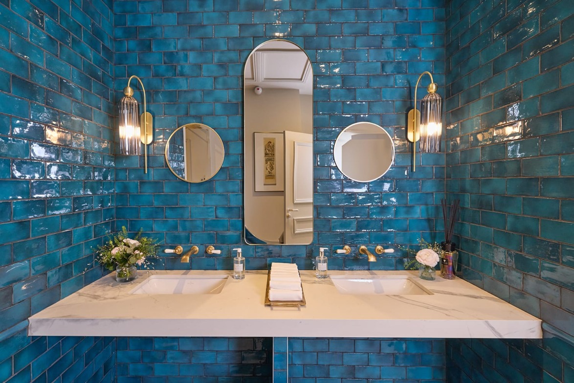 Modern bathroom with teal tiles, double sinks, oval mirrors, gold fixtures, and decorative plants.