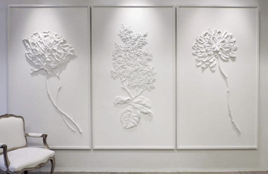 Three white floral relief sculptures on a wall, with an elegant chair positioned on the left side of the frame.