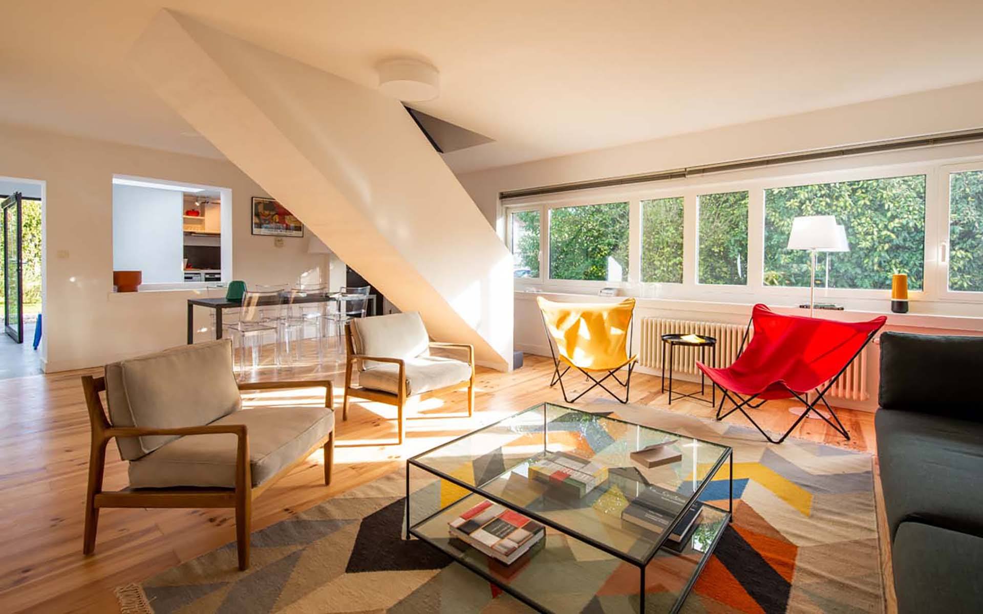 Modern living room with colorful chairs, glass coffee table, and large windows letting in natural light.