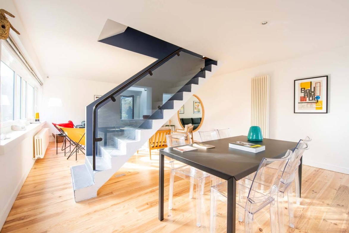 Modern living room with glass dining table, clear chairs, staircase, and bright natural light through large windows.
