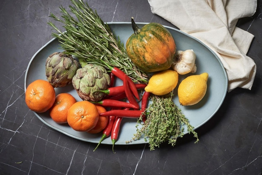 Plate with assorted vegetables and fruits including artichokes, oranges, lemons, chilis, pumpkin, garlic, and herbs on dark surface.