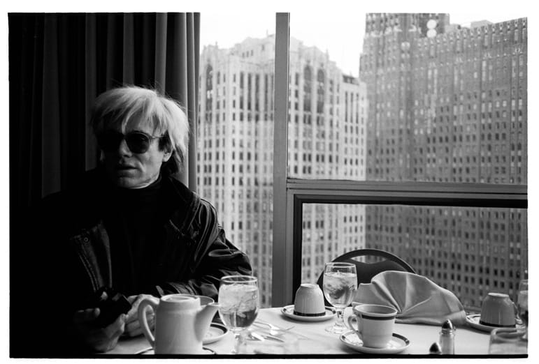 Person seated at a dining table with beverages, wearing sunglasses, in a high-rise cityscape restaurant setting.